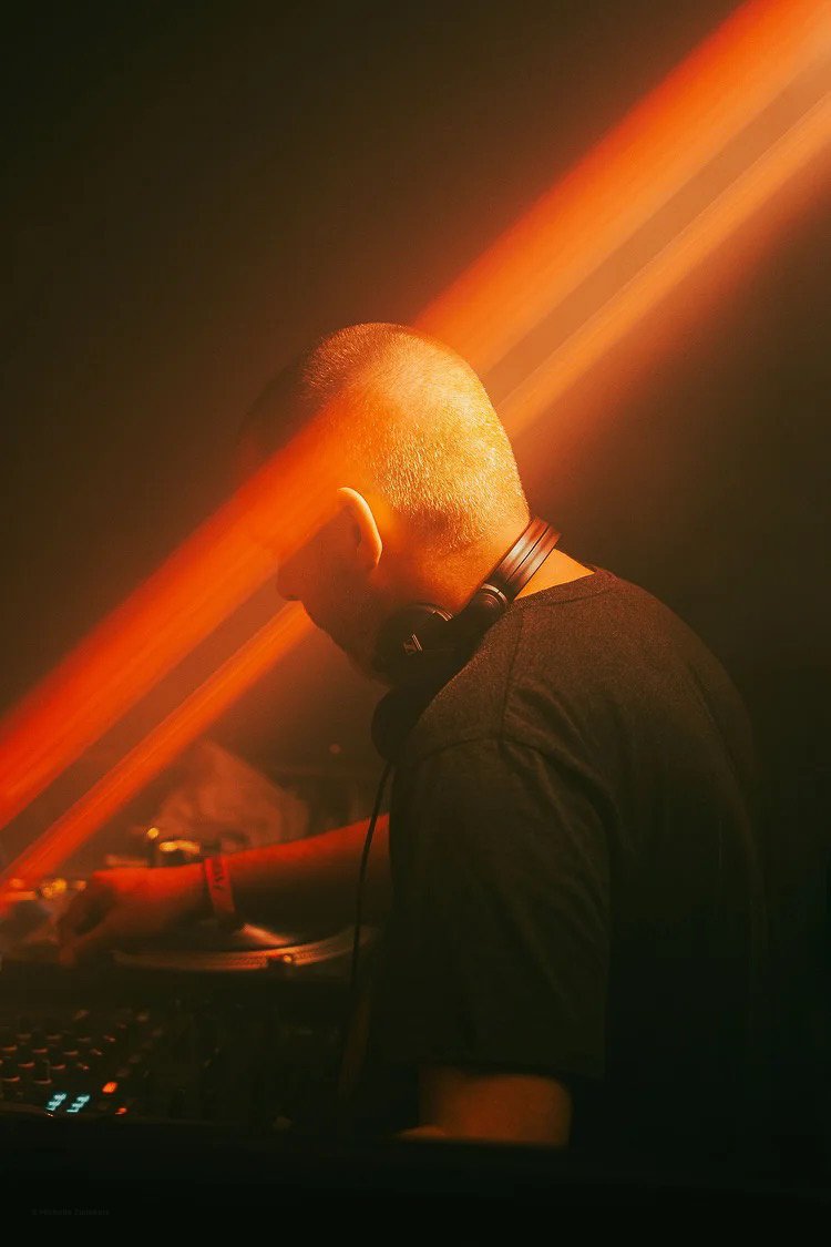 A DJ with a shaved head wearing headphones operating DJ equipment in a dark room with orange lighting.