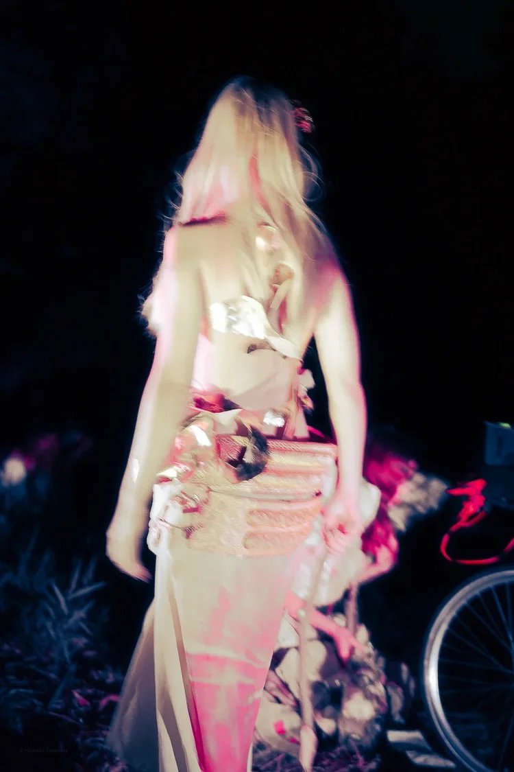 A person dressed in a beige or light-colored kimono standing outdoors at night, with a bicycle visible on the right side of the image.