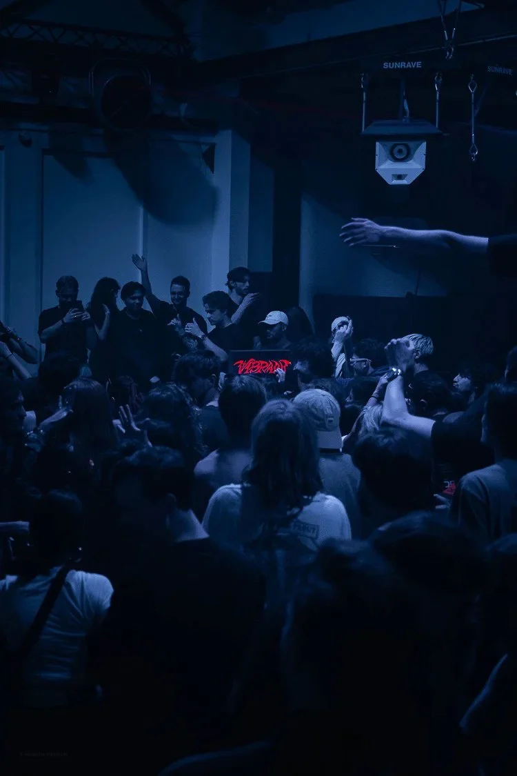 Crowd of people attending a concert or nightclub with blue lighting, some taking photos or videos, with a speaker and a hand reaching out from the right side.