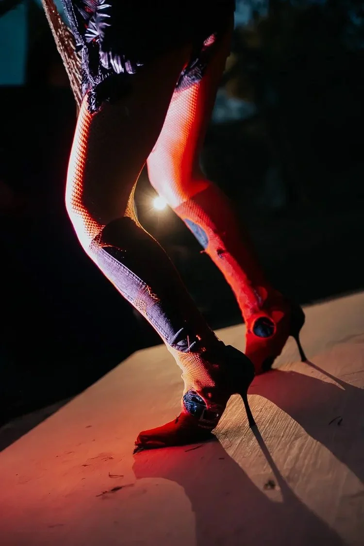 Close-up of a person in high heels and patterned leggings bending forward on a wooden surface with dramatic lighting and shadows.