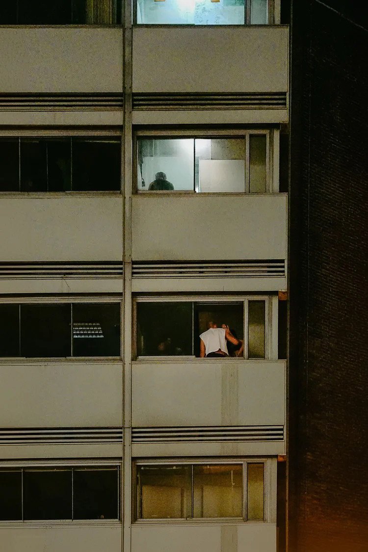 A person in a white shirt leaning out of an apartment window at night, with another person visible through a nearby window.