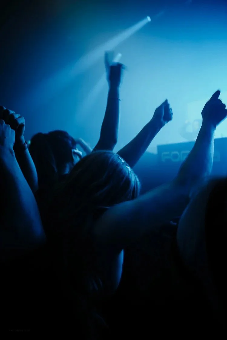 Crowd of people dancing at a club or concert with blue lighting, raising their hands in the air.