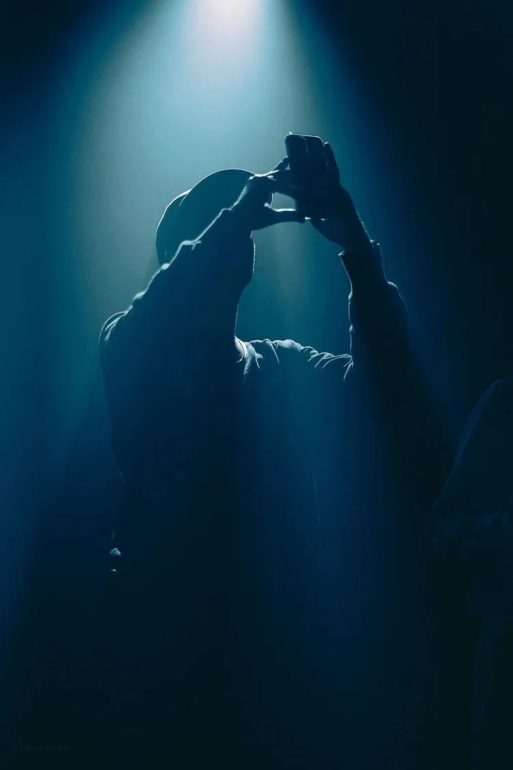Person taking a photo or selfie with a smartphone, backlit by a bright light in a dark room.