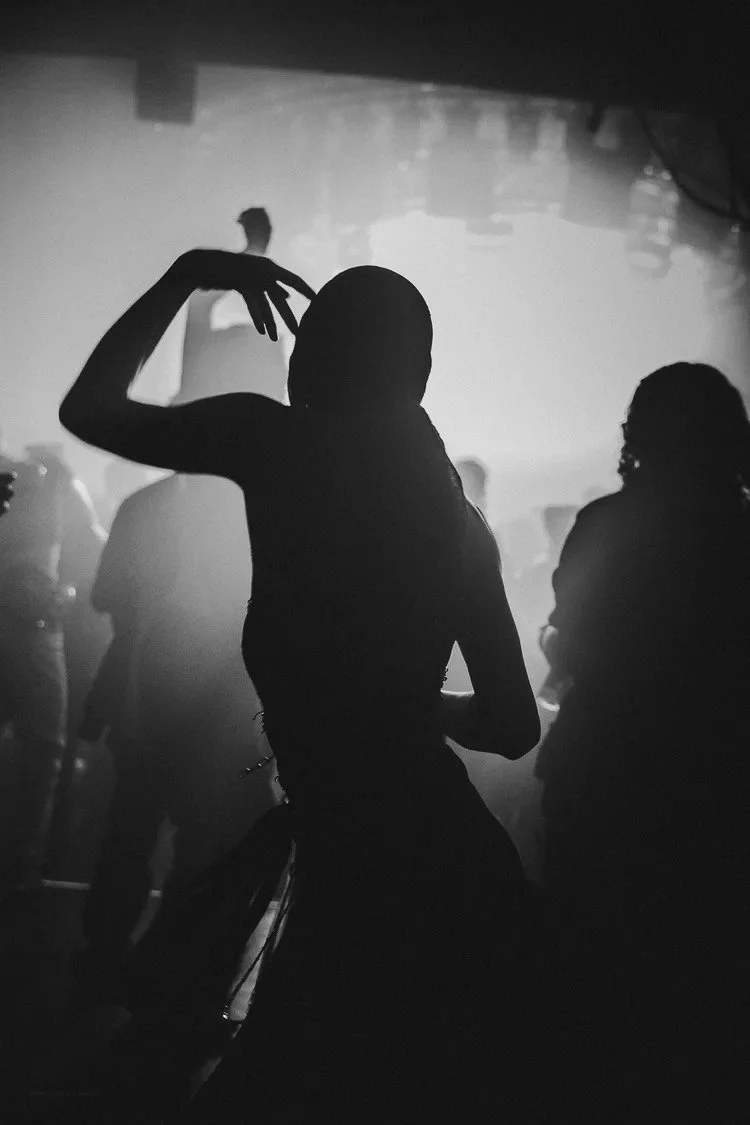 Silhouette of a woman dancing in a nightclub with other people in the background, illuminated by low lighting.