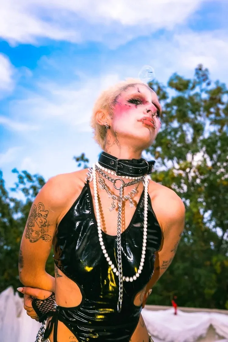 A person with short platinum hair, wearing heavy makeup with pink blush and lipstick, dressed in black shiny clothing with chains, pearls, and tattoos, outdoors against a background of blue sky and trees.