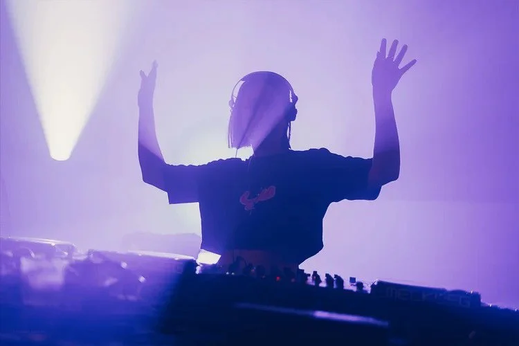 Silhouette of a DJ wearing headphones with hands raised, playing music in a purple-lit setting.