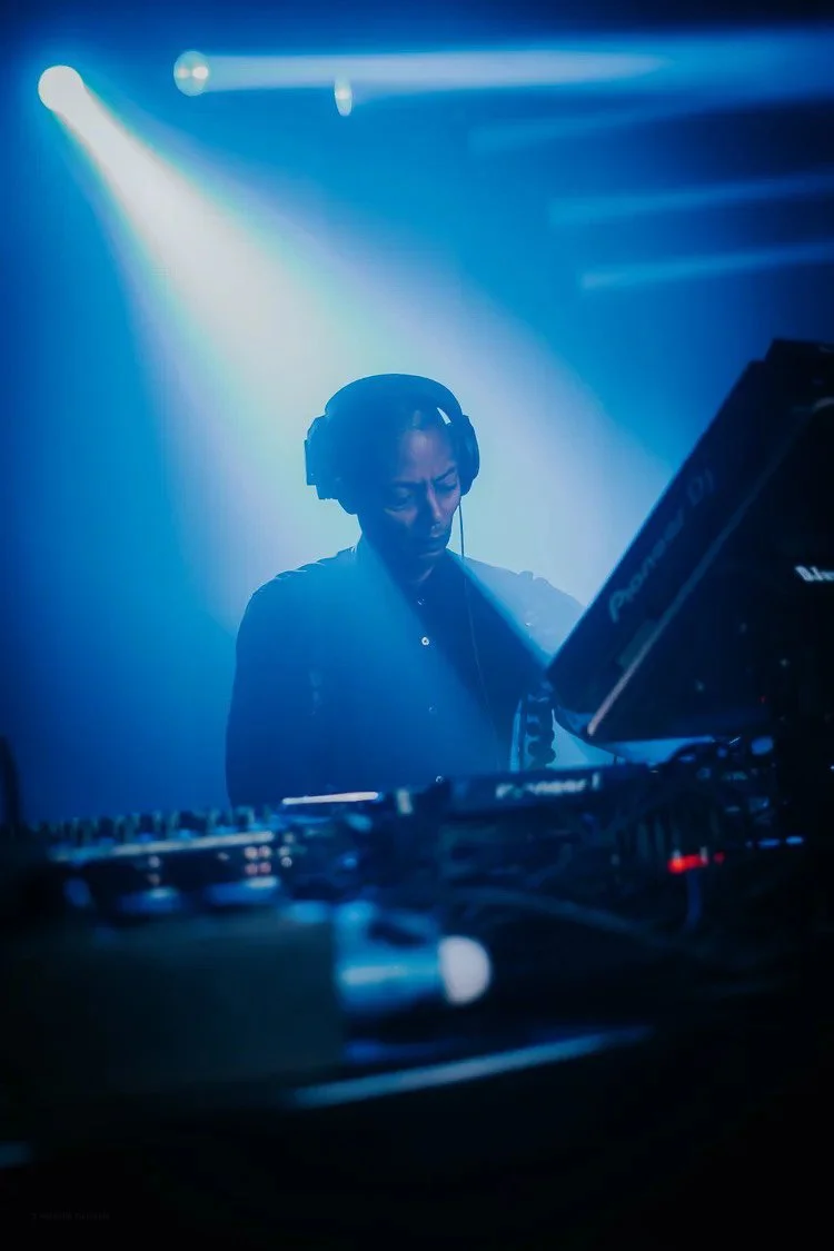 A DJ wearing headphones and a dark shirt performs behind a DJ mixer in a dimly lit venue with blue lighting and stage lights.