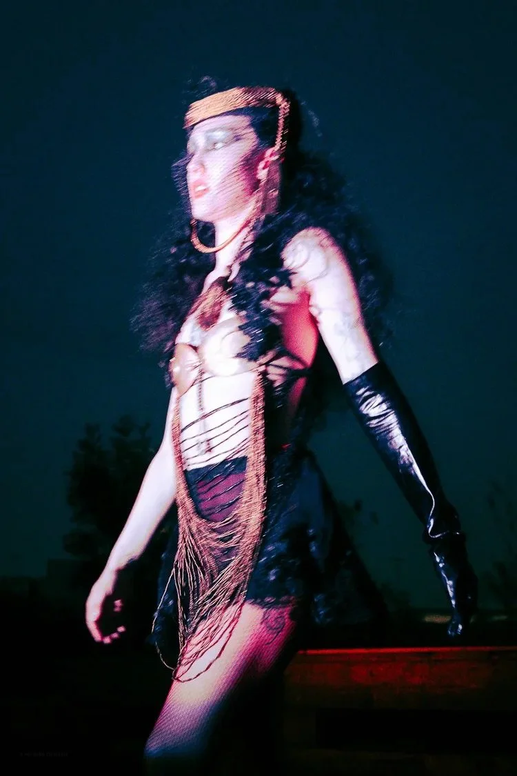A woman wearing a black, shredded dress with fringe details, long black gloves, and a headband with gold embellishments, standing outdoors at night.