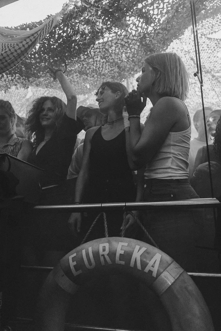 A group of women standing under a camouflage netting, with one woman holding a checkered cloth. There is a life preserver with the name 'EUREKA' hanging in the foreground.