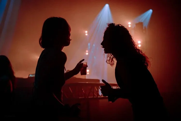 Silhouettes of two women talking and smiling in a dimly lit nightclub or concert venue with colorful stage lights in the background.