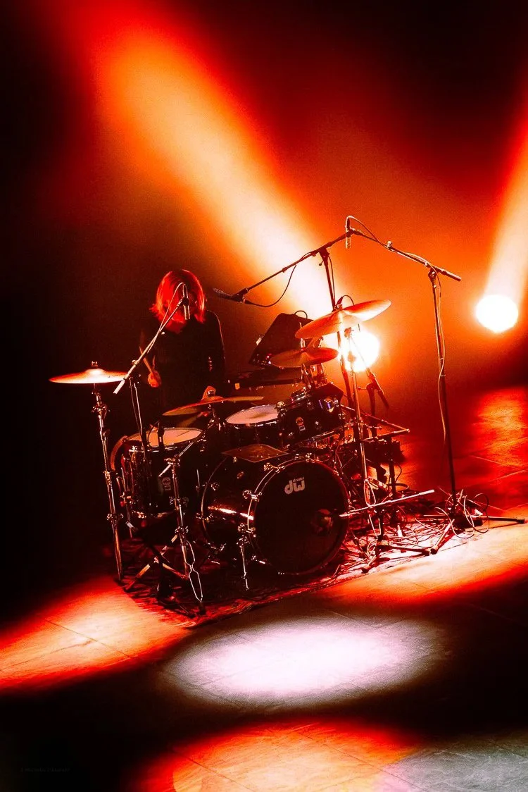 A musician playing drums on a stage with red and warm lighting, multiple microphones set up around the drum set.