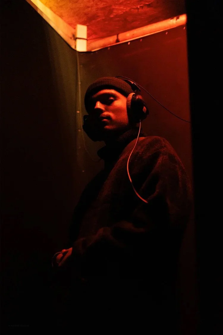 A person wearing a black beanie and headphones stands in a dark room with red lighting, looking towards the camera.