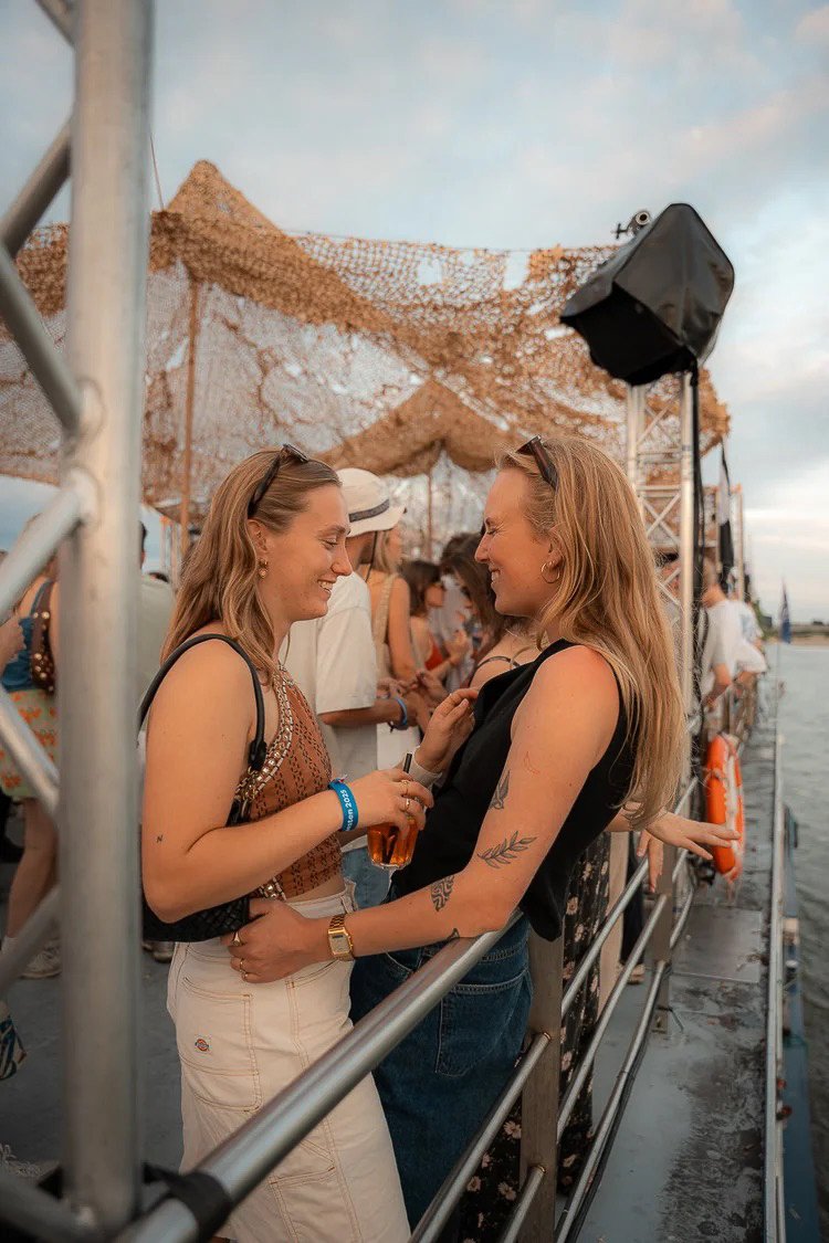 Two women with blonde hair smiling and holding drinks, standing close together on a boat during a sunny day with a background of other people and a decorated canopy overhead.