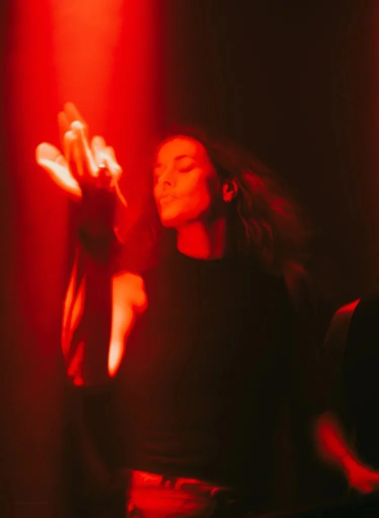 Woman with closed eyes and flowing hair in a dimly lit space illuminated with red light, standing with one arm raised and hand gesturing.
