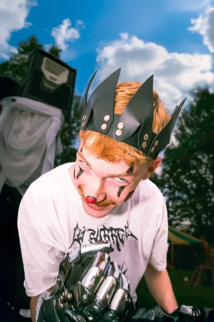 A person with red hair dressed as a clown, wearing a black crown with spikes, with face paint including a red nose, black lines, and a mustache, holding a metallic object, outdoors with a blue sky and clouds in the background.