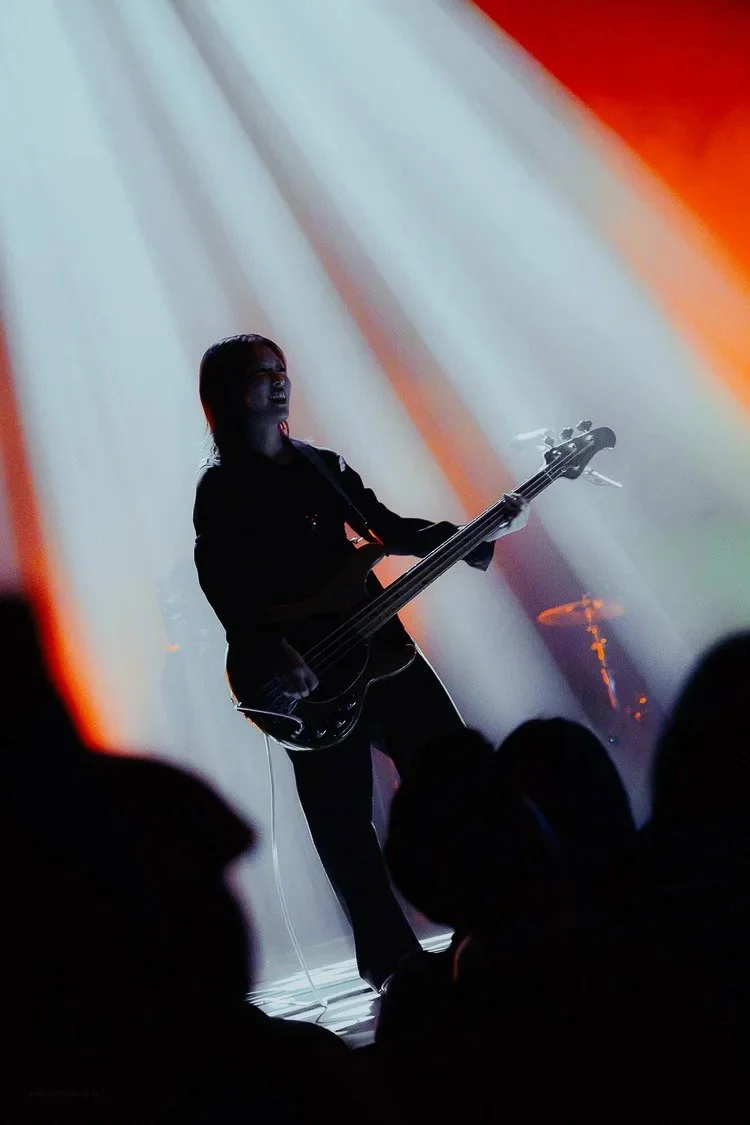 Silhouette of a musician playing an electric bass guitar on stage with dramatic lighting and orange-red background.