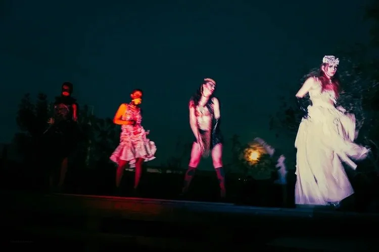 Four performers on a dark outdoor stage, dressed in theatrical costumes with dramatic makeup. The first on the left is in black with face paint, the second in a pink ruffled dress, the third in black lingerie with stockings, and the fourth in a white