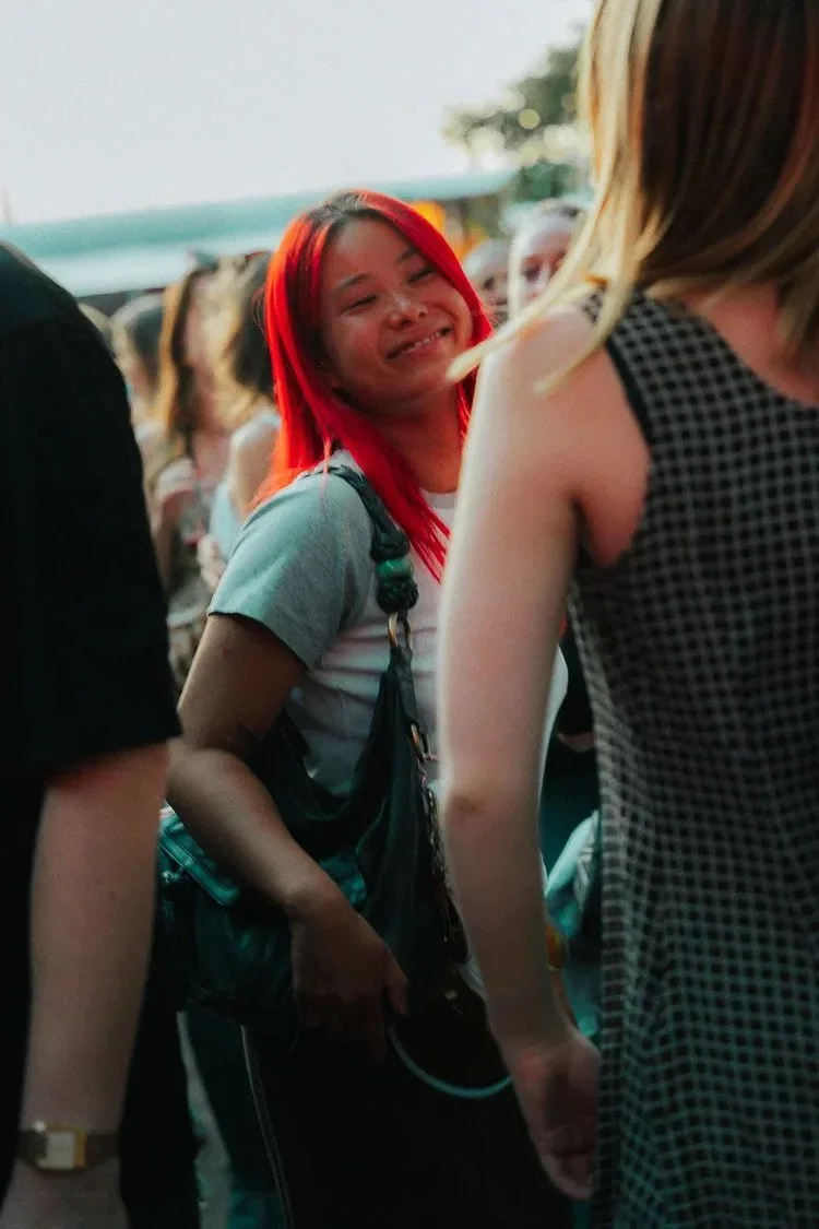 A young woman with bright red hair smiling and looking towards a person in a checkered dress at an outdoor event with other people in the background.