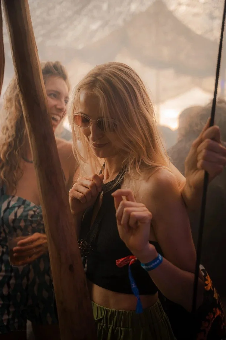 Two women at a party, one with curly blonde hair laughing and another with straight blonde hair, sunglasses, and a black crop top celebrating.