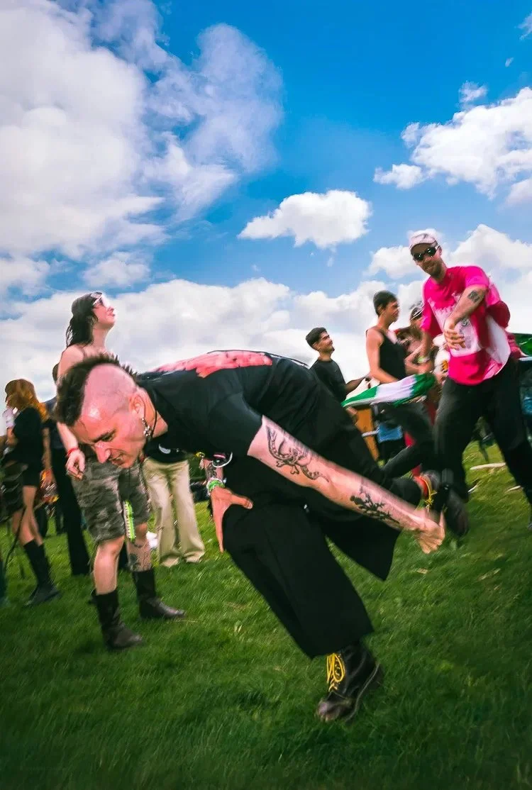 People at an outdoor event, some smiling and others engaged in activities, with a bright blue sky and scattered clouds overhead.