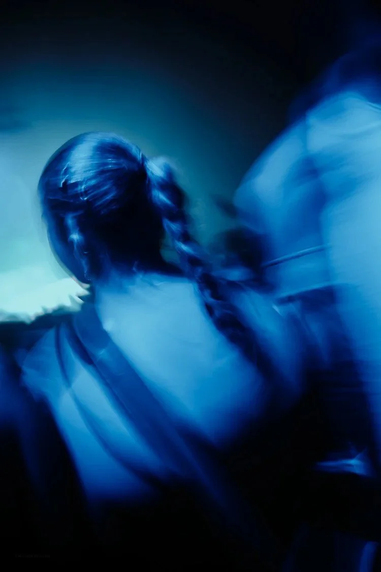 Blurred image of a person with a braid, illuminated by blue light, facing away in a dark environment.