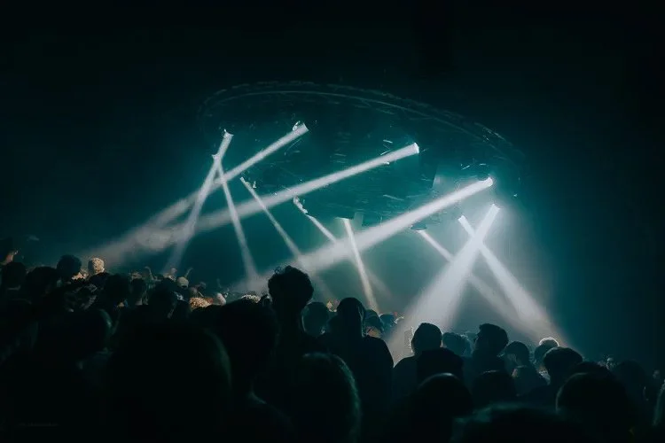 Crowd of people at a concert with bright stage lights and fog effects.