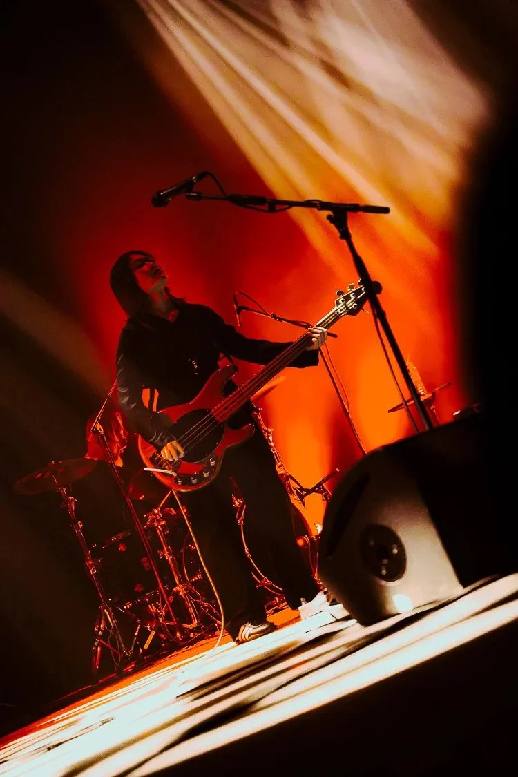 A musician playing an electric bass guitar on stage with orange and red lighting, standing near a drum set and microphone stand.