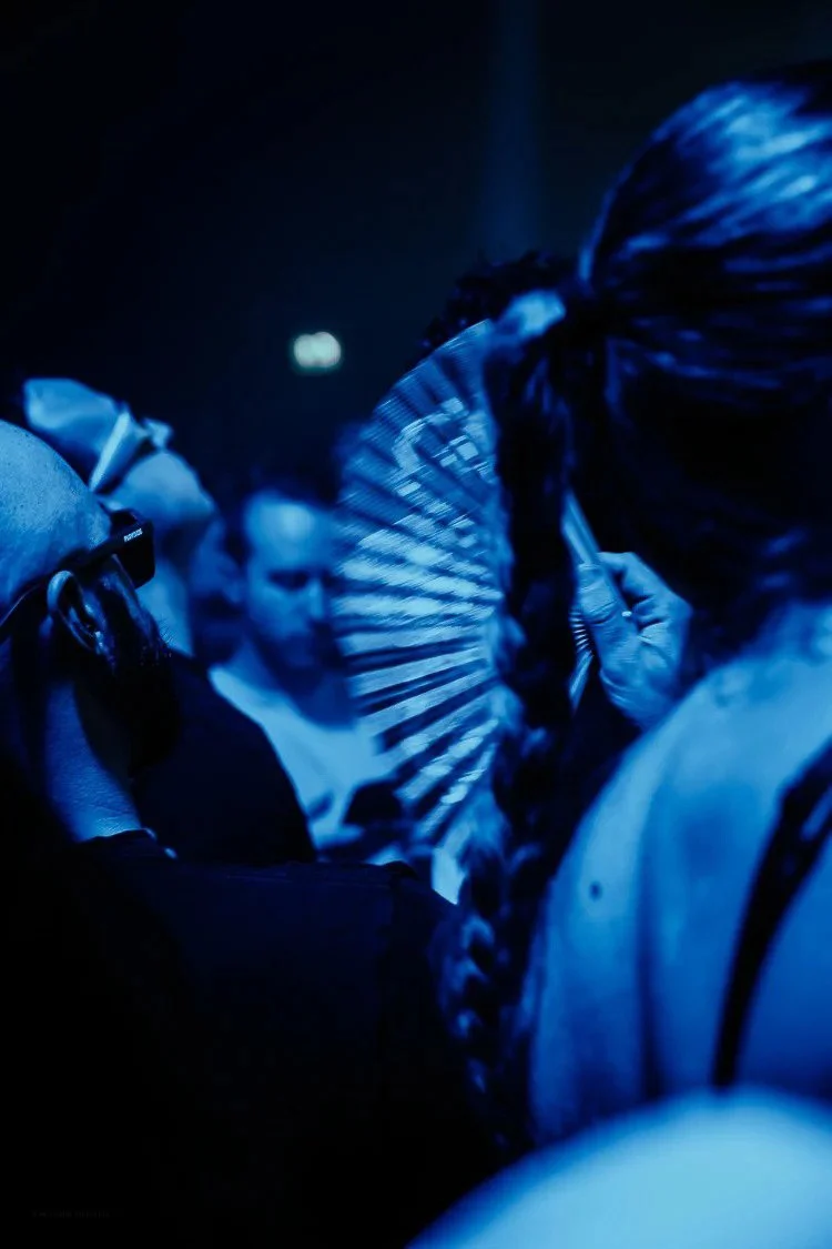 People getting makeup or face painting at an event, illuminated by blue lighting, with one person holding a fan.