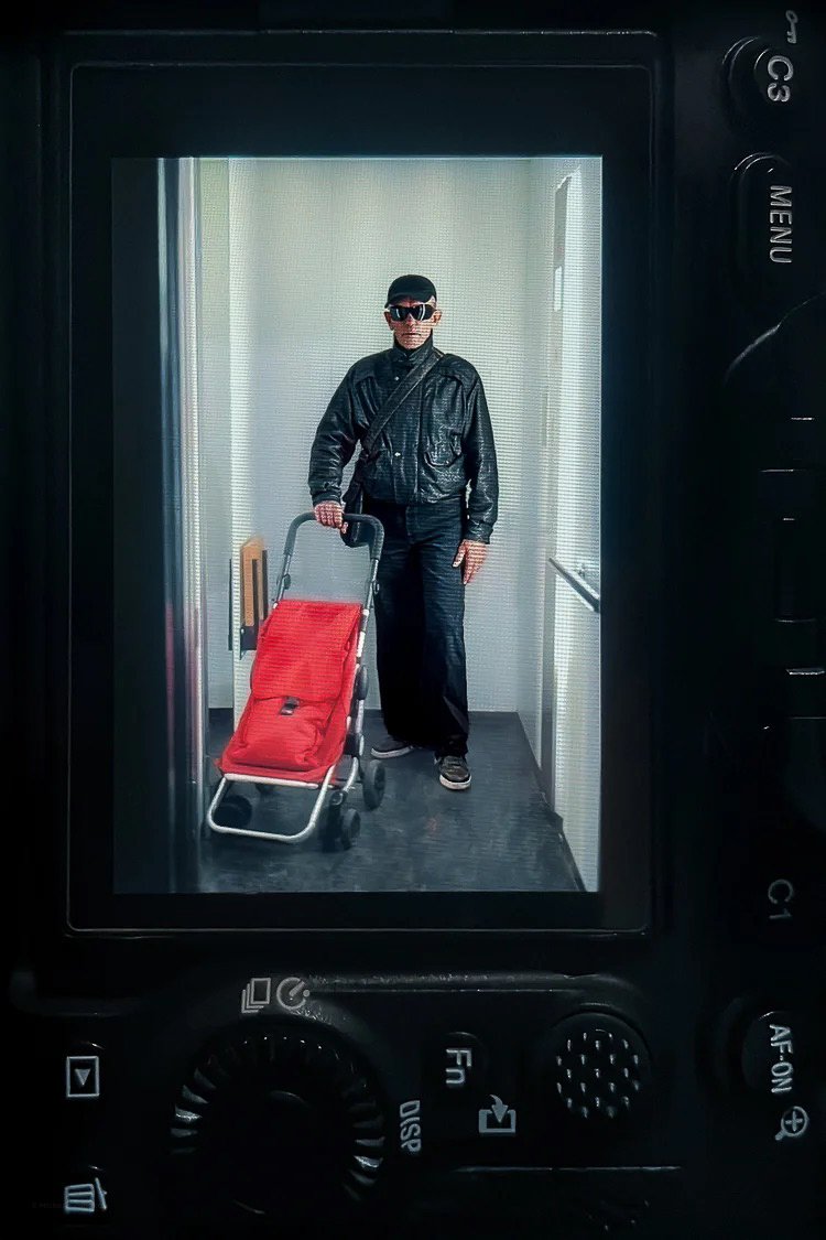 Person dressed in black leather jacket, black pants, black hat, and sunglasses standing in front of a mirror, pushing a red shopping cart in an elevator with white walls and black flooring.