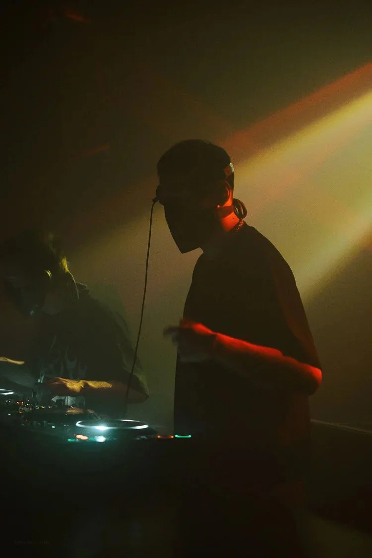 DJ wearing headphones, standing in a dark club with colored lights, adjusting music equipment.