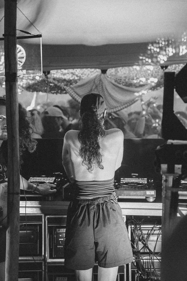 A woman with long hair, wearing glasses, a strapless top, and shorts, DJing at an outdoor event with a crowd in the background.