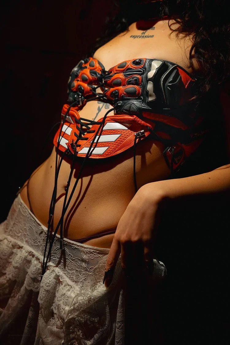 A woman wearing lace pants and a sports glove on her chest, with visible tattoos, in a dimly lit setting.