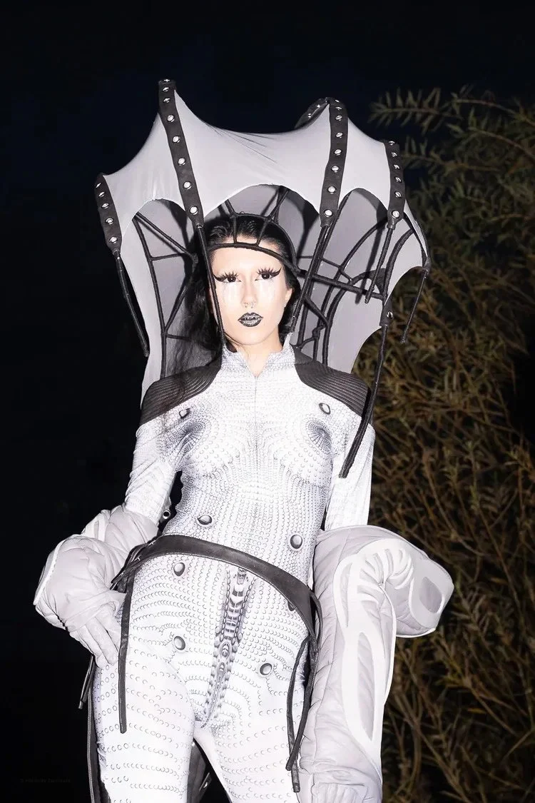 Person dressed in a futuristic, monochrome spider-themed costume with a large spider web umbrella, standing outdoors at night.