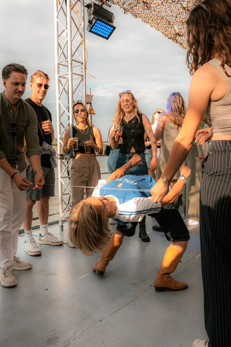 A group of people at an outdoor event dancing and socializing, with some holding drinks. One woman is doing a dance move while others watch and smile.