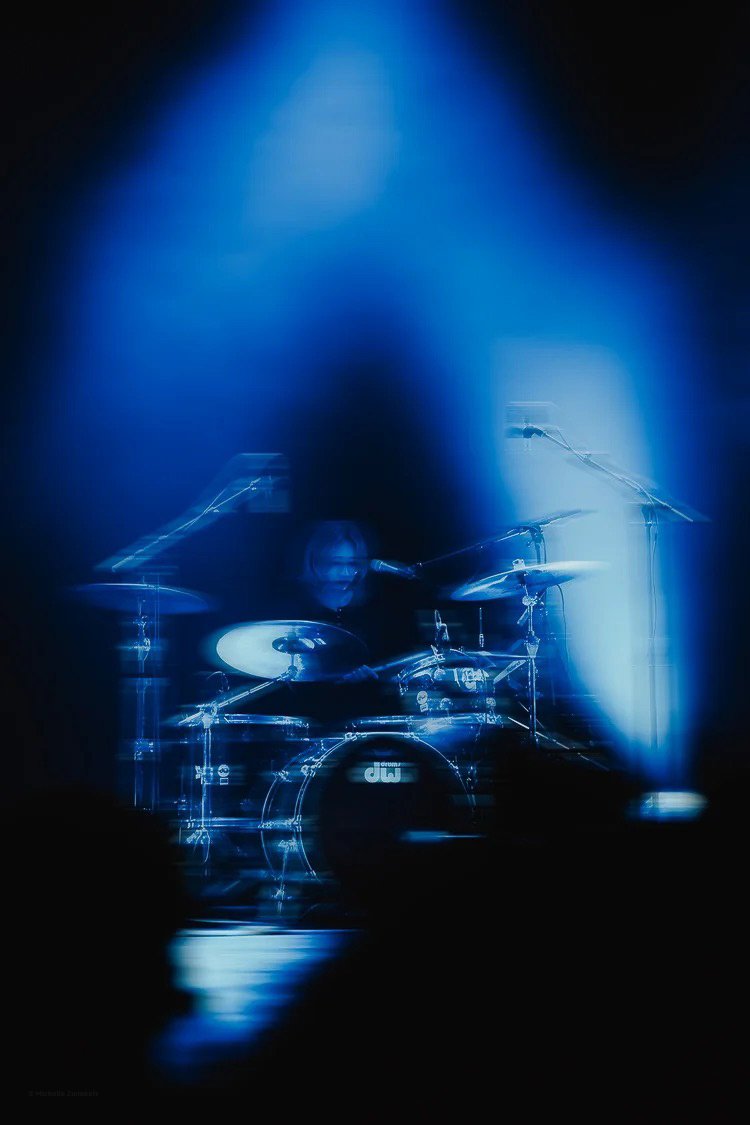 Musician playing a drum set on stage with dramatic blue lighting and fog, viewed through a blurred frame.