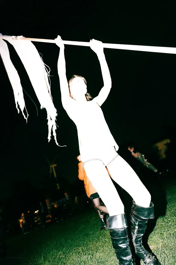 Person hanging from a horizontal pole at night, with shredded fabric hanging from the pole, and other people in the background outdoors.