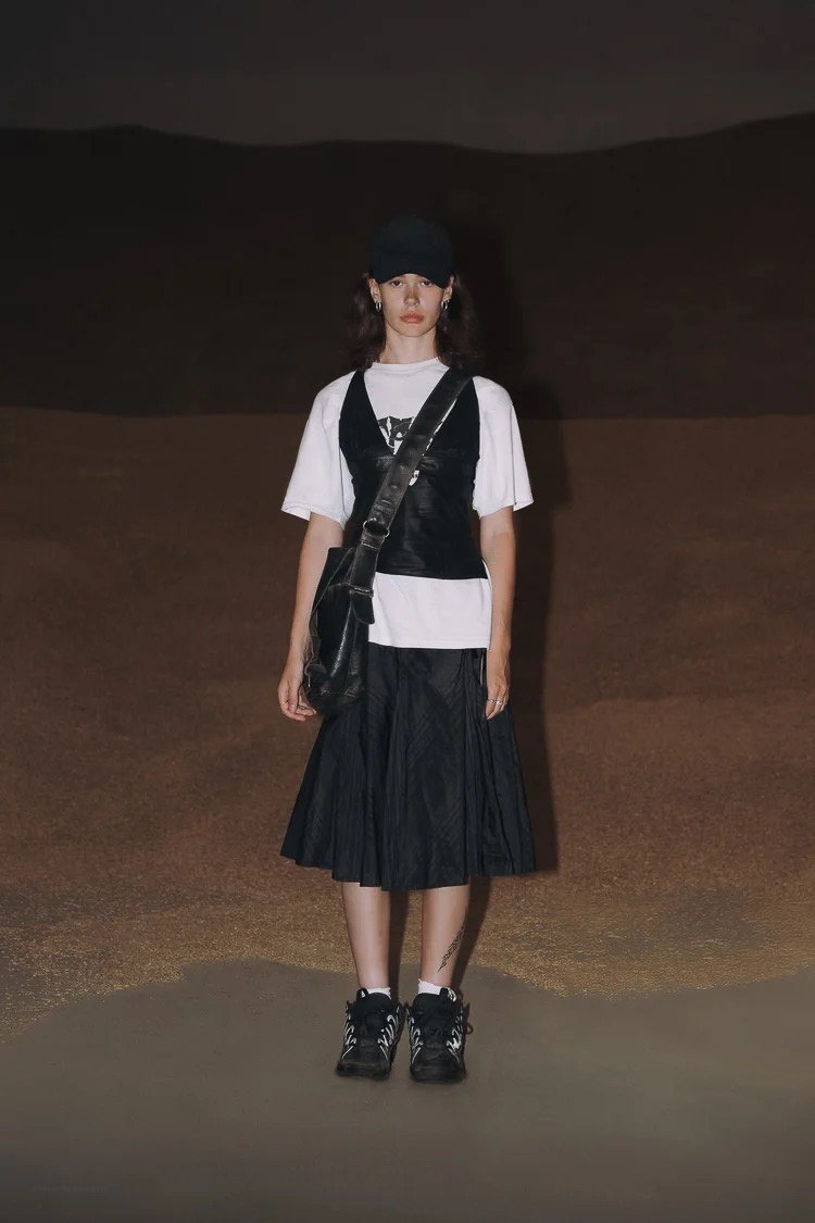 Young woman standing in a desert, wearing a black cap, white T-shirt, black vest, black pleated skirt, and sneakers, with a black crossbody bag.