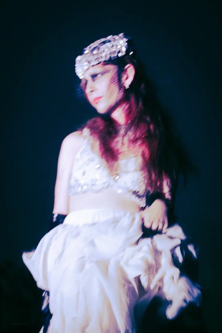 Person in a sparkling headpiece wearing a white dress with ruffles, holding the fabric of the dress, with dark red hair against a dark background.