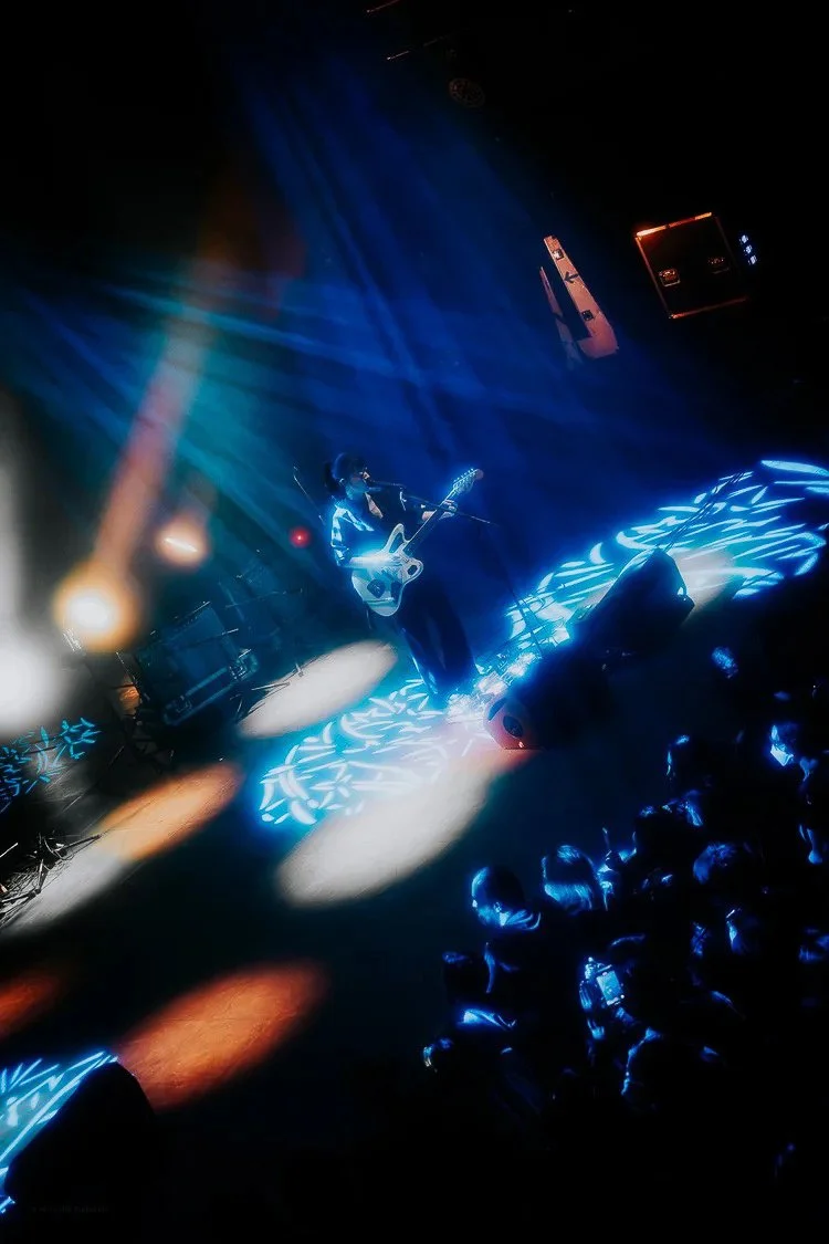 A musician performing on stage with a guitar surrounded by blue and orange stage lights, and a live audience watching.