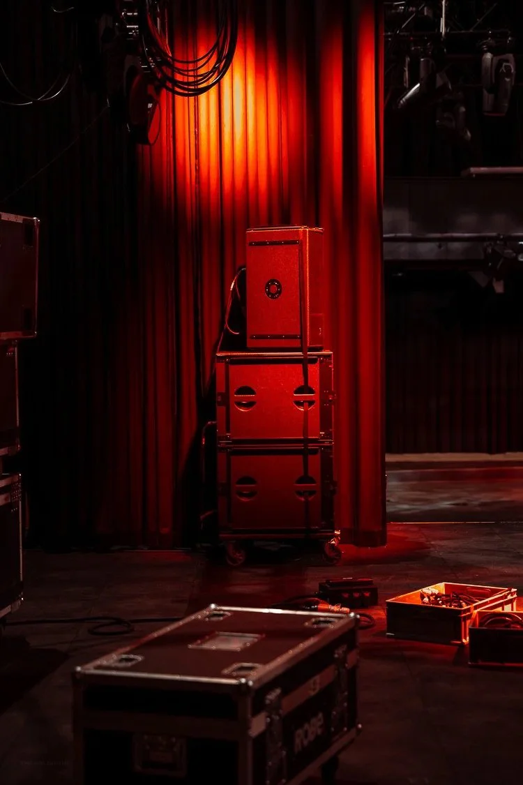 Stage with red curtains, stacked speaker cabinets on a cart, and equipment cases on the floor.