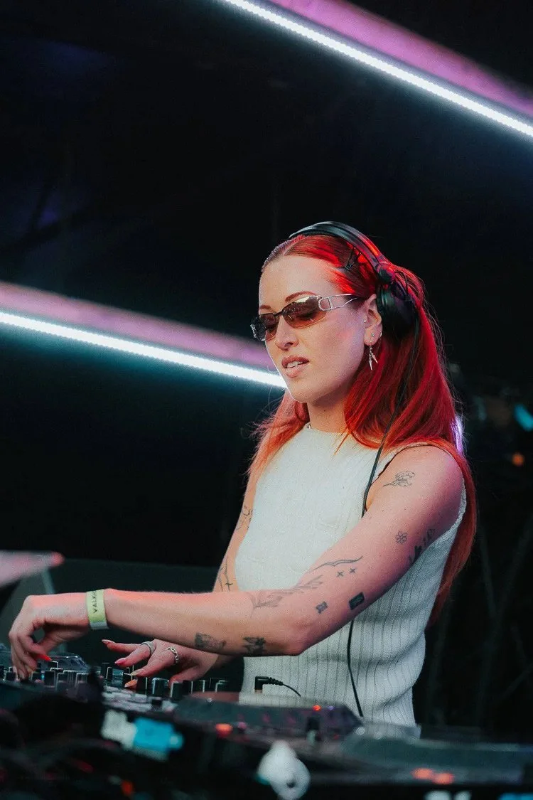 A female DJ with long red hair, tattoos on her arms, wearing sunglasses, headphones, and a sleeveless white top, is mixing music at a nightclub with colorful LED lights in the background.