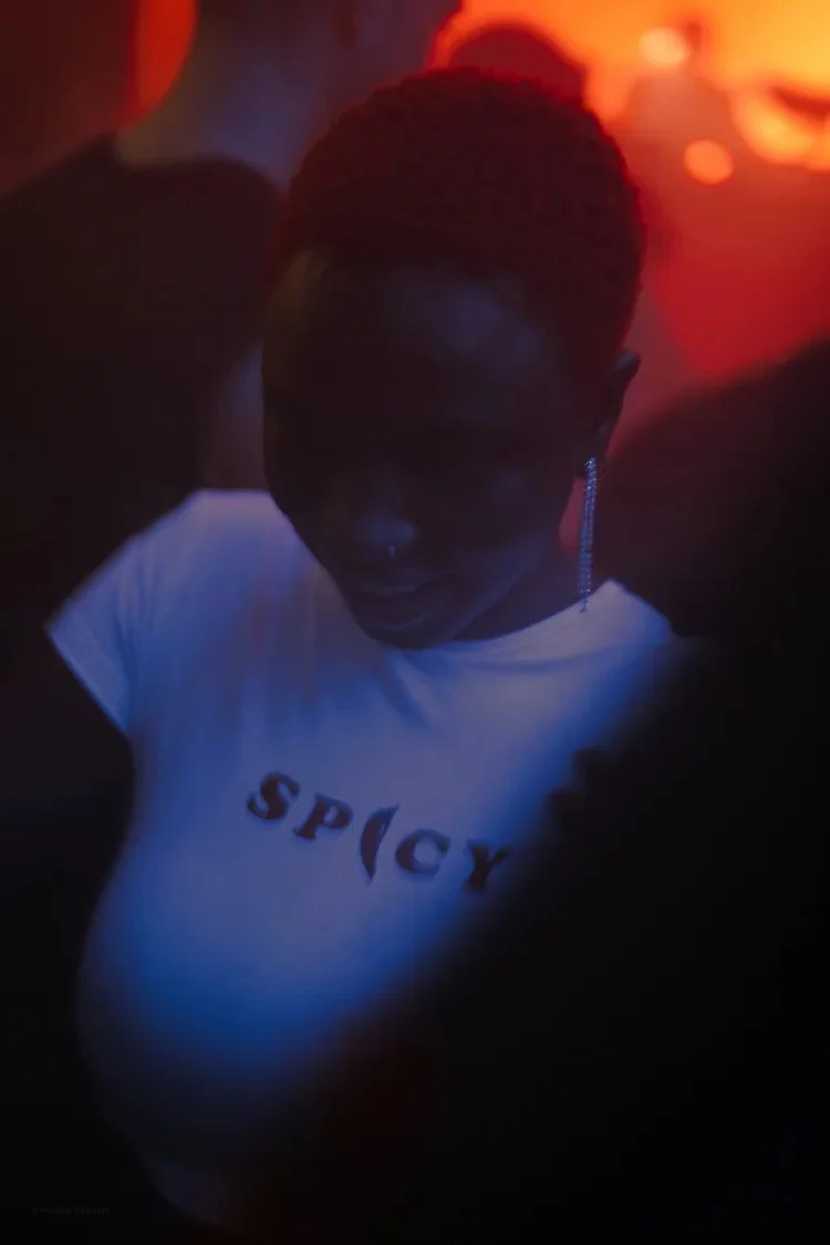 A woman in a white shirt with partially visible red text, wearing earrings, dancing in a dark setting with warm orange lighting.