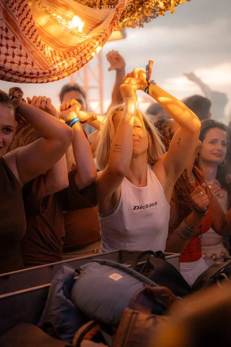 A woman at an outdoor concert or festival raising her arms with a joyful expression, surrounded by other people, under a warm sunset sky.