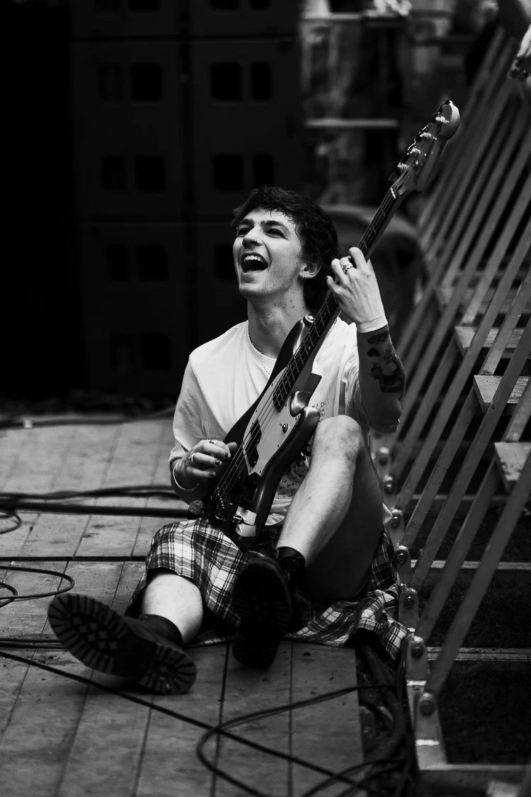 A young man sitting on a wooden floor with a guitar, smiling and laughing, in an indoor setting with musical equipment around.