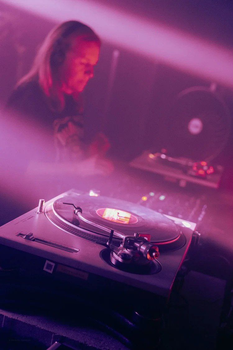 A DJ with long hair in a dimly lit, pink-purple environment, focusing on a turntable and mixer.