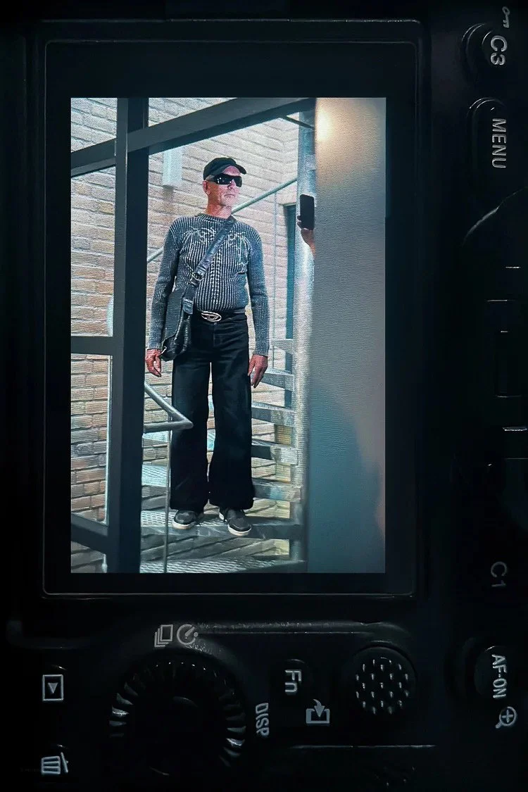 A person taking a mirror selfie outdoors, standing on spiral stairs near a brick building, wearing dark pants, a sweater, sunglasses, a cap, and carrying a bag.