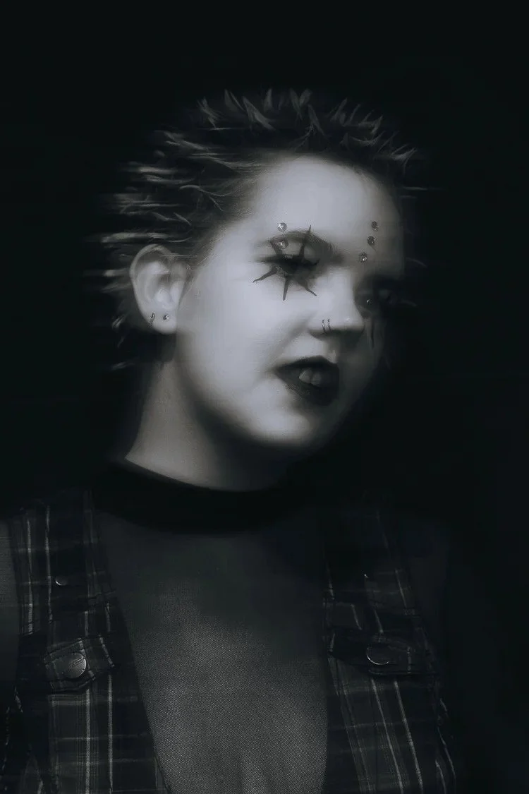 A person with short spiky hair, wearing dark makeup and gothic clothing, looking to the side with a dark background.