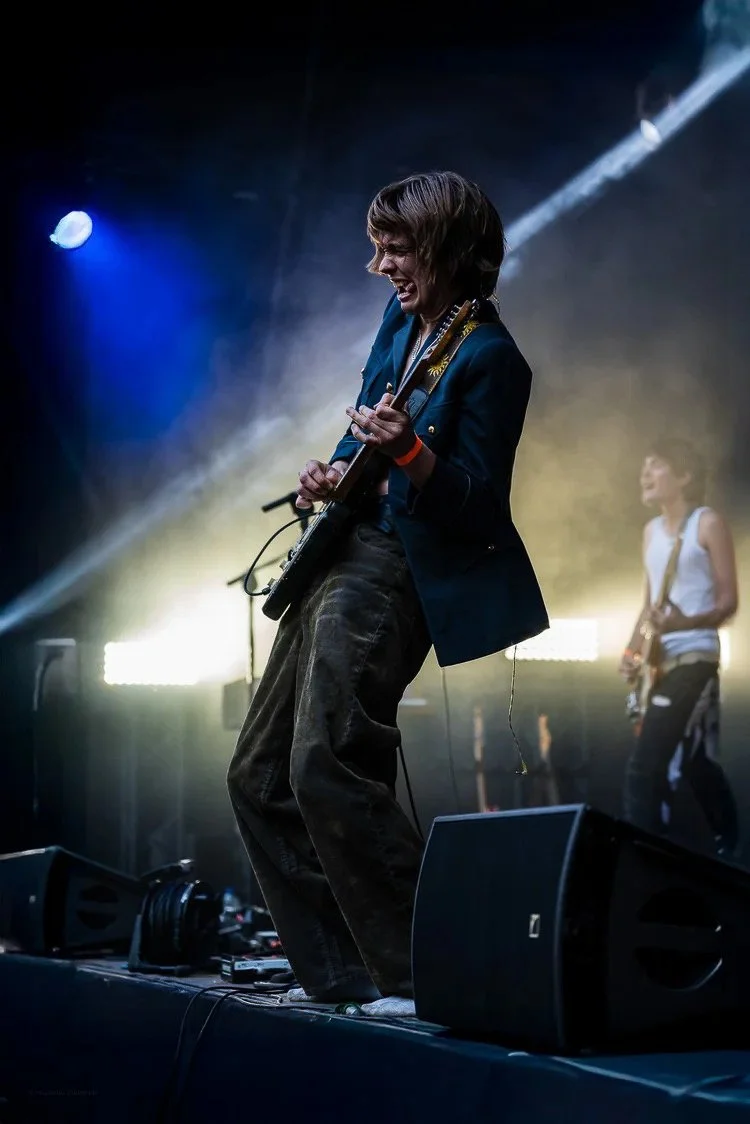 Female musician passionately playing electric guitar on stage with colorful stage lights and another band member in the background.