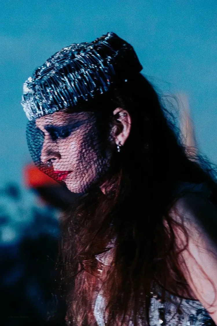 A woman wearing a shiny, textured headpiece with black netting over her face, red lipstick, and earrings, with long dark hair and a blurred background.