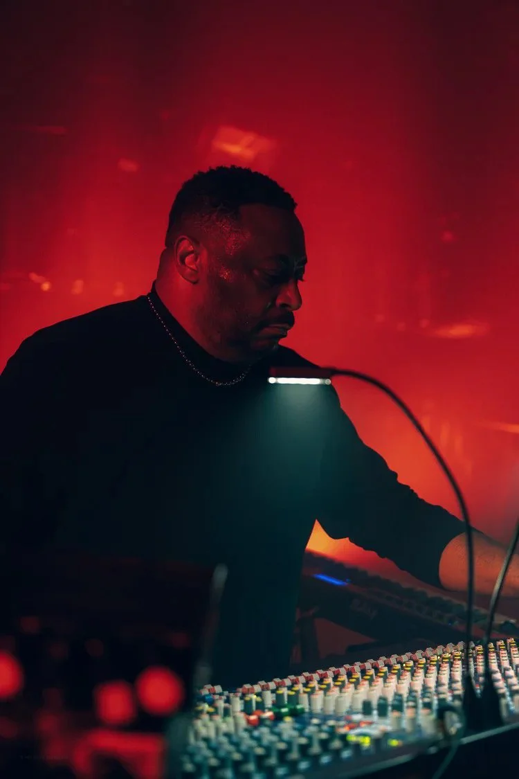 A man with short dark hair and a dark shirt is operating sound mixing equipment with colorful knobs, in a dimly lit environment with red lighting.